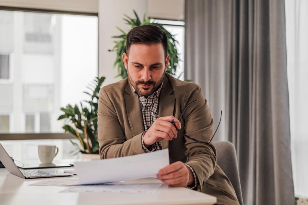 Man dressed in a suit sits at a desk and thoughtfully looks at a paper with a pen in his hand